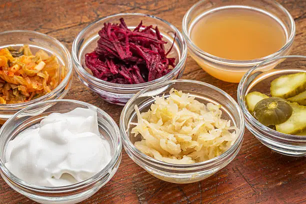 a sampler of fermented food great for gut health - glass bowls against wood:  kimchi, red beets, apple cider vinegar, coconut milk yogurt, cucumber pickles, sauerkraut