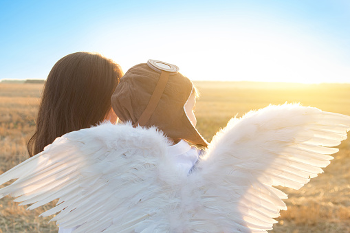 Boy and his mother playing the aviator and look on sunset. The boy in a pilot hat and angel wings sitting on hands of his mother.