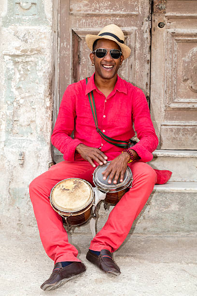 Professional Cuban Conga Drum Player A well known professional Cuban conga drum player percussionist and the afro-cuban Santeria priest in red clothes and Panama hat sitting in front of an old door, playing bongo drums, smiling, Old Havana, Cuba. salsa music photos stock pictures, royalty-free photos & images