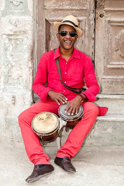 A well known professional Cuban conga drum player percussionist and the afro-cuban Santeria priest in red clothes and Panama hat sitting in front of an old door, playing bongo drums, smiling, Old Havana, Cuba.