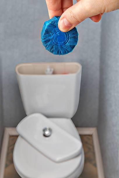 Close-up, blue cleaning tablet for sanitizing a toilet bowl, for flush mechanism inside cistern of toilet, in hand housewife, against the backdrop of household toilets.