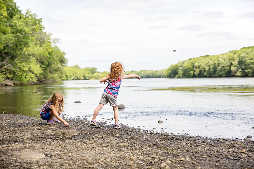 Two young girls (sisters) playing on the bank of the Mississippi River on a summer day in Minnesota, USA. The younger sister has just thrown a rock towards the water - the rock is visible in the air above the trees near the right side of this image. The older sister is crouched on the riverbank picking up stones to throw into the water.