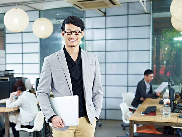 portrait of young asian entrepreneur young asian entrepreneur standing in office with laptop computer under arm. founder stock pictures, royalty-free photos & images