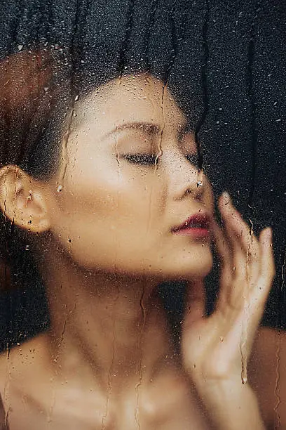 Face of beautiful Asian young woman behind wet glass