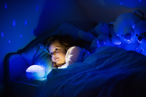 Little girl reading a book in bed. Dark bedroom with night light projecting stars on room ceiling. Kids nursery and bedding. Children read before bedtime. Toddler child playing with lamp and bear toy.