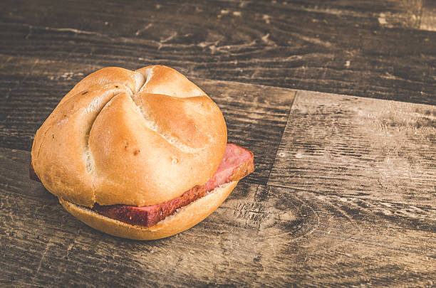 A professional DSLR photo of a fresh and very tasty leberkaese semmel on a wooden oak kitchen table. High angle viewpoint. The leberkaese is a traditional Austrian and German Bavarian food. It is a baked bread roll with hot pork meat and cheese. Lovely details in this image. Photo made in a modern photo studio with professional light. Soft vintage post processing, for a lovely retro look.