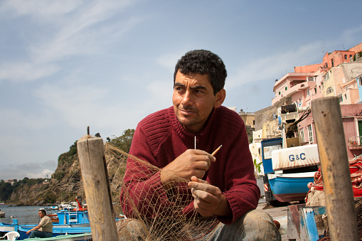 Procida, Italy - May 16, 2008: A fisherman repairing a fishing net on the island of Procida in the Bay of Naples, Italy, with small fishing boats,  cliffside homes, and sky in the background.