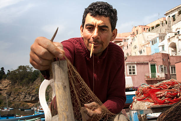 Procida, Italy - May 16, 2008: A fisherman repairing a fishing net on the island of Procida in the Bay of Naples, Italy. Small fishing boats,  cliffside homes, and sky in the background.