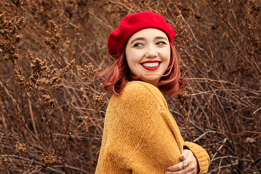 Beautiful young woman enjoying autumn wind in nature. She has red hair, red lipstick, and is wearing a red beret and a yellow wool sweater. Selective focus on her face, blurred drying plants in the background. Head and shoulders outdoors horizontal shot with copy space. This was taken in Montreal, Canada.