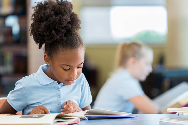 Cute African American schoolgirl reading a book in the library Adorable African American elementary age schoolgirl sitting at a table in the library and reading a book. Cute young African American girl reading a book at the school library. school uniform stock pictures, royalty-free photos & images