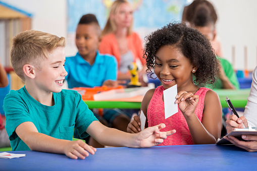 Cute Caucasian male elementary age student using flashcards with adorable African American female student. Students using flashcards in class together, sitting at a desk in a elementary school classroom.