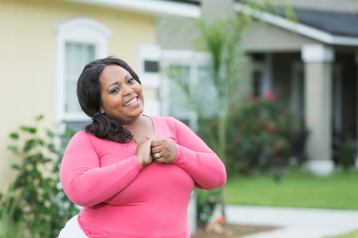 A young, overweight African American woman standing in the front yard outside her house, smiling at the camera. She is wearing a pink shirt.