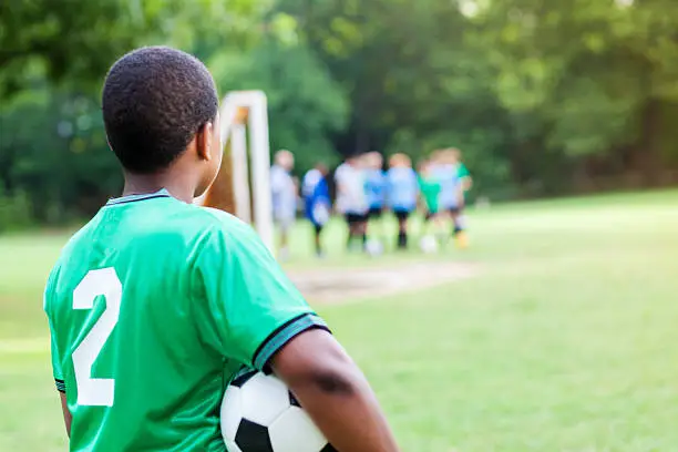 Preteen African American boy stands on the sidelines waiting to play in the soccer game. He watches his teammates play the game. He is holding a soccer ball. He is wearing a green jersey with the number 2 on the jersey. The back of the boy is seen in the photo.