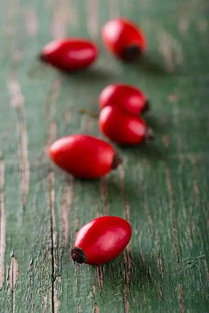 Vertical photo of several red hips with detail of the top of first fruit. Hips are placed in a row on wooden board with worn green color.