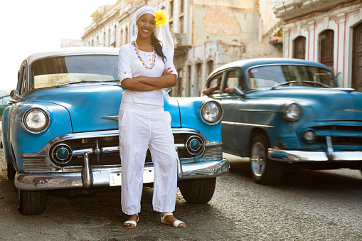Beautiful, young, smiling, Cuban woman standing in front of a restored, blue, vintage American car from the 60s on the street in Old Havana, Cuba. A woman wearing a Santeria white dress. Another old blue car driving past. Santeria religion combining aspects of West African religion with christianity. Low angle view