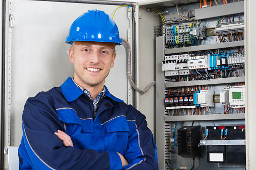 Portrait Of A Happy Young Male Electrician Standing In Front Of Fusebox