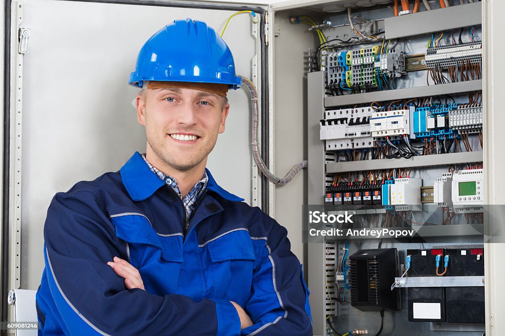 Portrait Of A Happy Young Male Electrician Portrait Of A Happy Young Male Electrician Standing In Front Of Fusebox Electrician Stock Photo