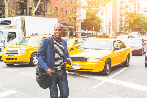 Black man crossing a street in New York. Young man wearing smart casual clothes commuting in the city, with buildings and cars on background. Commuter lifestyle and youth culture concepts.