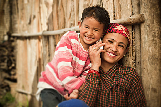 Happy Asian mother and daughter on the phone. Outdoor day time image of mature Nepalese women attending a phone call while her daughter trying to listen by putting her ear on phone at the same time and leaning over the shoulder of her mother. They are sitting with the wall of their traditional wooden hut. Two people, waist up, horizontal composition with copy space. central asian ethnicity stock pictures, royalty-free photos & images