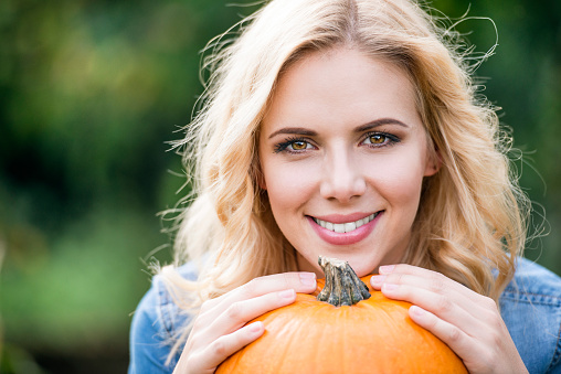Close up of beautiful young blond woman in denim shirt with orange pumpkin. Autumn nature.