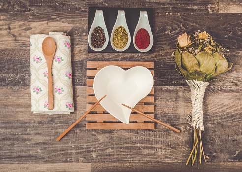 A professional DSLR concept stock photograph of a table ready laid with many lovely details. Delicious mouth watering and aromatic mixed seasoning, a wooden spoon, asian chopsticks and a heart shaped bowl on a wooden bambus plate. There is a dried flower bouquet and a beautiful white and pink floral napkin. This stock image is a concept for mealtime. Professional photo studio shot with high quality photo equipment. Vintage post processing for a lovely retro effect.