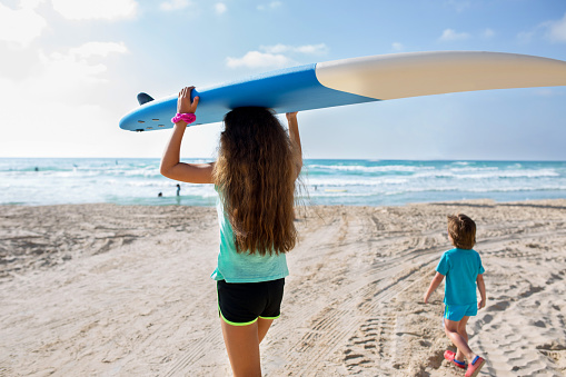 Rear view of teenage girl carrying surfboard on her head while walking on the beach with her little brother.