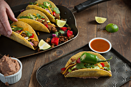 A high angle close up photograph of a hand reaching for one of three beef tacos  sitting in a griddle pan. There is also another taco and a small bowl of spicy salsa sitting on a tray in the foreground.