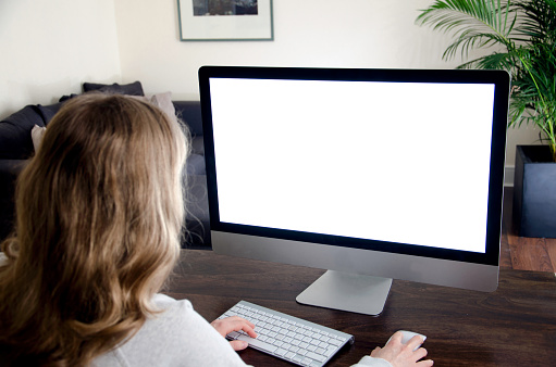 A blond young woman is sitting on a desk and is working on a computer with blank monitor screen left white for your own copy space of message. The photo was taken from behind over the models shoulder in a domestic room. 