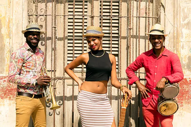 Cuban musical band, the trio consisting of a well known musicians standing against the wall. Beautiful young woman standing in the middle, holding a guitar. The man on the left holding the small drums bongos, and a musician on the left holding a trumpet. Havana, Cuba