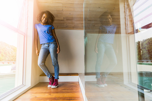 A photo of confident young woman leaning on wall by window. Full length of beautiful female is wearing casuals. She is standing at home.