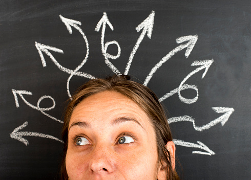 A forehead of the woman who is pondering about possible solutions of the complicated problem. Many arrows with different directions are drawn around her head. Black chalkboard background.