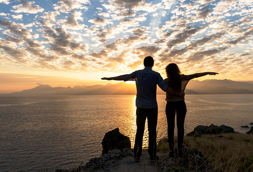 Portrait of couple standing with arms outstretched on cliff at sunset