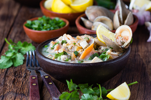 Latin American food. Seafood shellfish ceviche raw cold soup salad of seafood shellfish almejas, lemon, cilantro onion in clay bowl on wooden background. Traditional dish of Peru or Chile