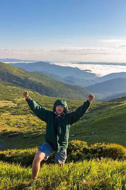 istock Young man standing on top of mountain, holds hands up 609818314