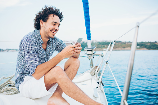 Smiling young man traveling on a yacht in the sea in the summer and checking messages.