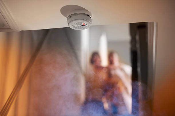 smoke alarm smoke rises up towards a second floor in a two two storey house . Upstairs we can see a parent carrying a small child and running from a bedroom as they have been woken by the smoke alarm which is shown in focus at the edge of the ceiling leading to the stairwell . fire alarm smoke detector smoke danger stock pictures, royalty-free photos & images