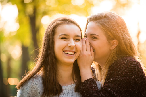 Teenage girl whispering secret to her friend - outdoor in nature at sunset