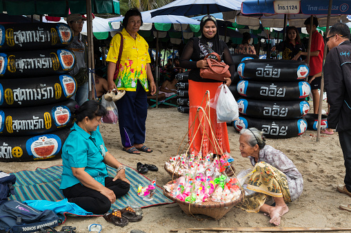 Bang Saen, Thailand - January 10, 2015: Very old Thai woman selling toys on the beach, Bang Saen, Thailand. Thai tourists are watching her. In the background car tubes for swimming. Bang Saen is a popular destination for Bangkok people during vacations