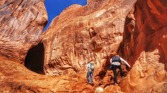 Male and female hikers walk inside one of Arches National Park's many alcoves inside the Fiery Furnace backcountry maze of a hike. Slot cave ahead.