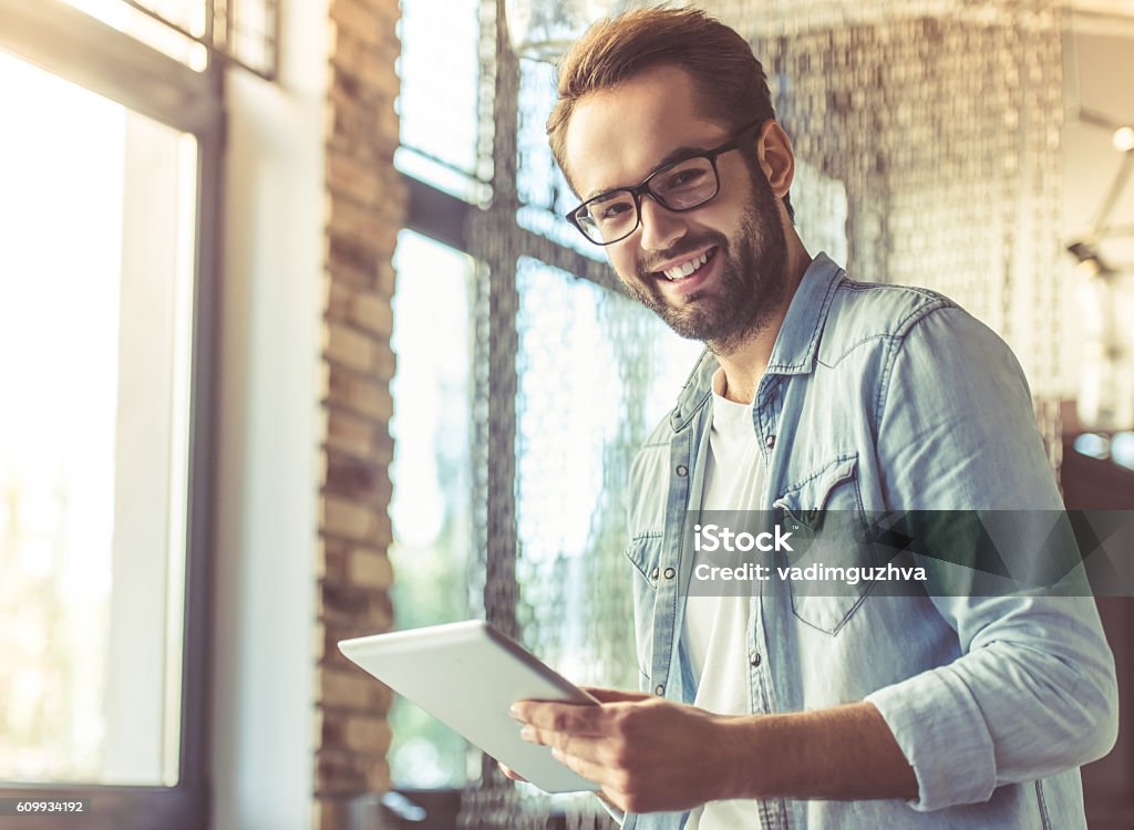 Handsome businessman working Handsome businessman in casual wear and eyeglasses is using a digital tablet, looking at camera and smiling while standing in the office Adult Stock Photo