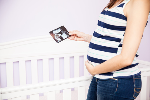 Closeup of a young pregnant woman touching her belly and looking at an ultrasound picture while standing next to a crib