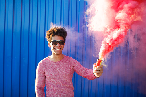 Portrait of a young black hipster man smiling and holding a pink smoke bomb over a blue color background. Youth and lifestyle concept