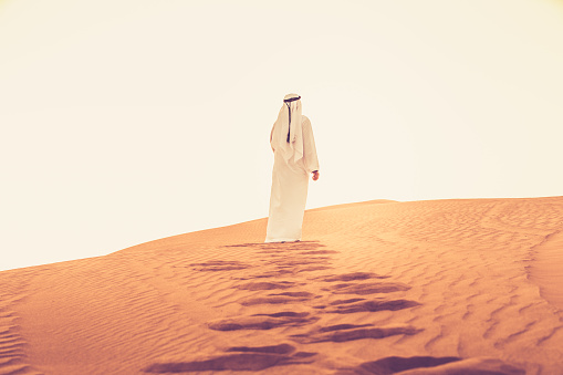 Arab walking on the sand dunes of Dubai and looking at the bright sunlight.