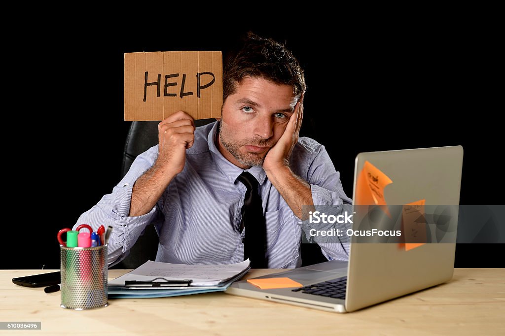 tired desperate businessman in office stress working asking for help young desperate businessman suffering stress working at office computer desk holding sign asking for help looking tired exhausted and overwhelmed by heavy work load isolated on black background Frustration Stock Photo