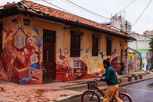 Bogota, Colombia - July 20, 2016: A cyclist hurriedly crosses the street, in the historic La Candelaria district of the Andean capital city of Bogota in the South American country of Colombia, quite unmindful of the the street art that decorates the building across the road. He has an Arhuaca Mochila strung across his shoulder. His movement causes a blur, but enhances a sense of urgency. The style of architecture is typical Spanish Colonial: tall doors and barred windows. The roof has terracotta tiles. Photo shot in the morning sunlight, on an overcast day. Horizontal format.