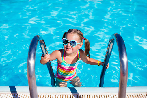 Happy laughing little girl playing in outdoor swimming pool on a hot summer day. Kid in colorful bathing suit and goggles learning to swim in tropical resort. Water fun for children.