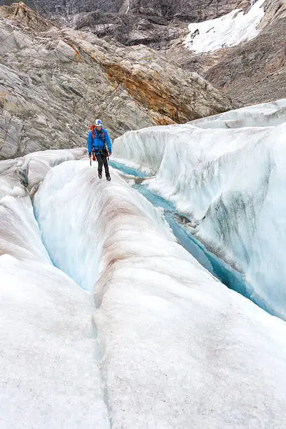Man in blue jacket, helmet and backpack walks along a narrow ridge between crevasses with the safety of crampons attached to his boots on a trekking adventure on Lemon Glacier, Juneau Icefield, Juneau, Alaska, USA