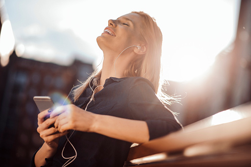 Young woman sitting on a bench, holding smartphone with headphones and listening to music