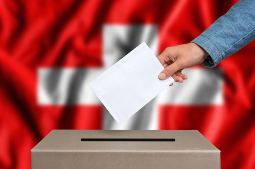 Election in Switzerland. The hand of woman putting her vote in the ballot box. Swiss flag on background.