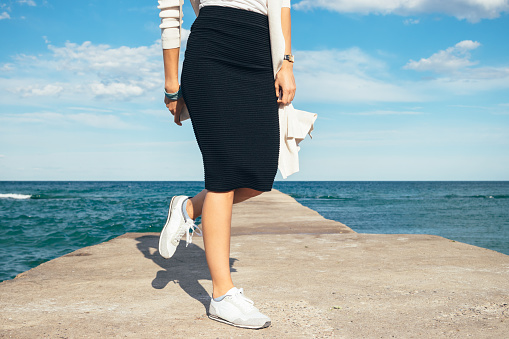 Details of women's clothing: Female legs in a skirt and sneakers on the background of the sea